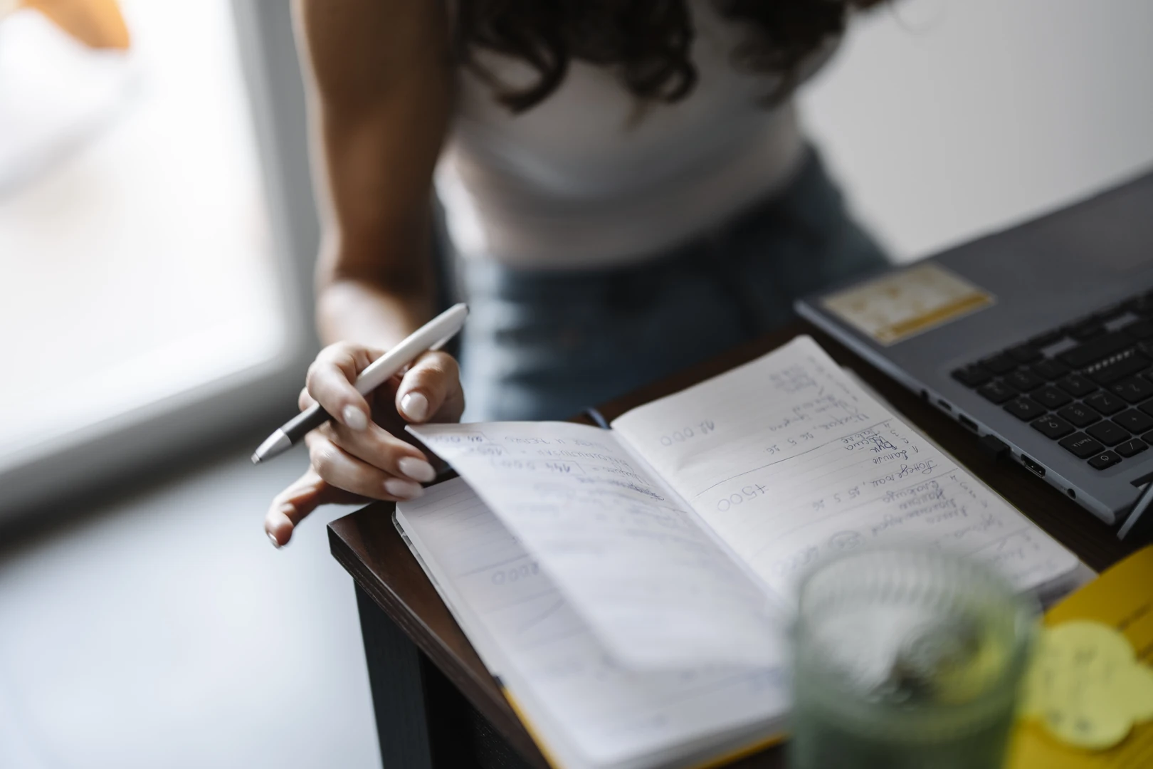 A person writing notes next to a laptop and a glass of water